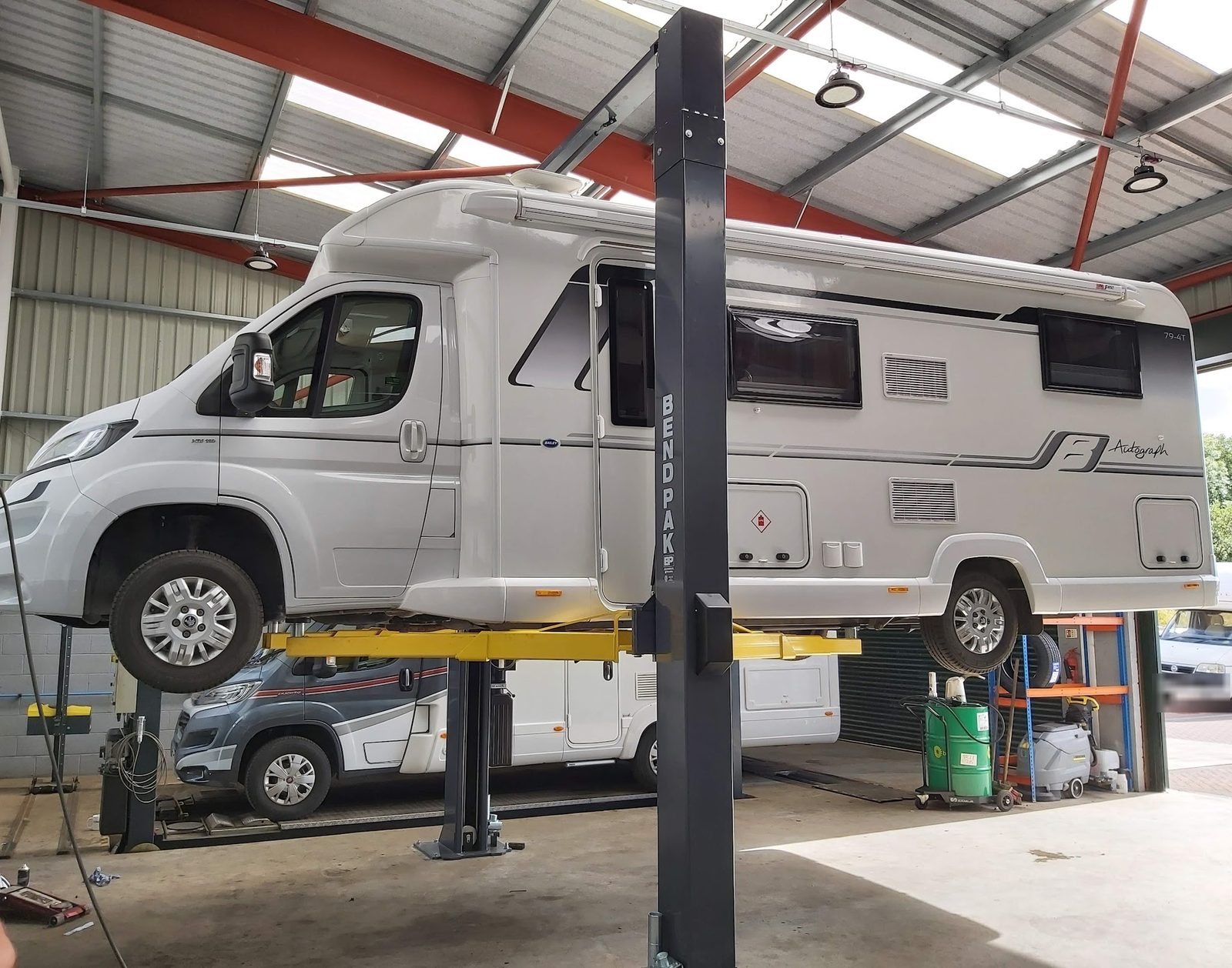 A Bailey motorhome raised on a two-post lift in the workshop, with a second motorhome parked on the ramp behind