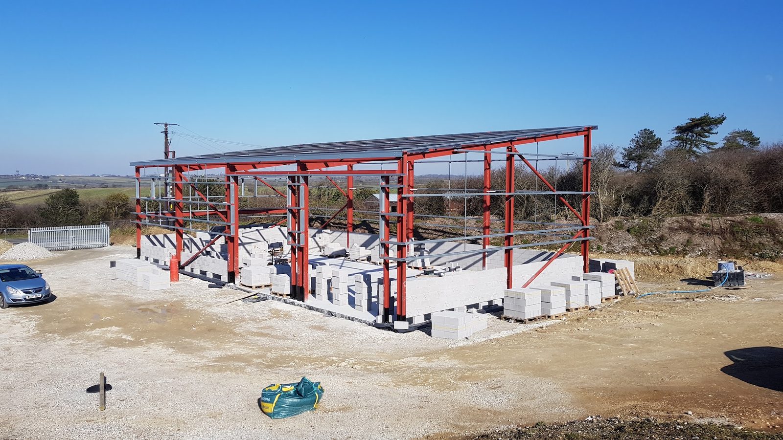 Red steel frame and block foundations of the new workshop at Quintrell Downs, open fields behind